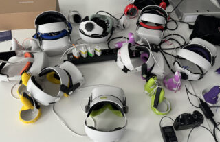 Auf einem Tisch sind mehrere VR-Headsets mit verschiedenfarbigen Kopfhörerabdeckungen und Controllern angeordnet und an eine Steckdosenleiste und Ladekabel angeschlossen.