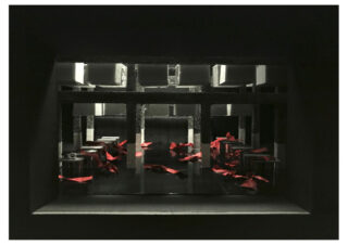 Ein dunkler, kastenförmiger Raum mit mehreren in Reihen angeordneten Tischen und Stühlen, einige davon mit roten Bändern oder Stoffen bedeckt.