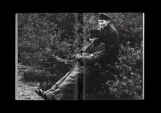 Ein Schwarzweißfoto von zwei Personen im Freien; eine sitzt in Mantel und Hut an Büschen, die andere steht in Uniform und Mütze dahinter, beide blicken geradeaus.