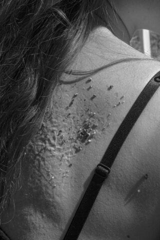 Eine Nahaufnahme der Schulter einer Person mit langen Haaren, auf deren Haut Schmutz und Partikel zu sehen sind, neben einem schwarzen BH-Träger.