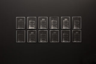 Zwölf gerahmte abstrakte Drucke mit dunklen, rechteckigen Formen sind in zwei Reihen vor einer schwarzen Wand angeordnet.