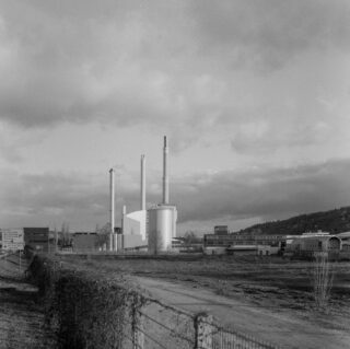 Ein Schwarzweißfoto einer Industrieanlage mit drei hohen Schornsteinen, umgeben von Gebäuden und offenem Land unter einem bewölkten Himmel.