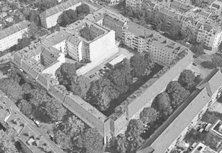 Luftaufnahme eines rechteckigen Apartmentkomplexes mit einem Innenhof voller Bäume und geparkter Autos, umgeben von Stadtstraßen und anderen Gebäuden.