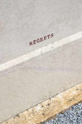 Auf einer hellen Betonwand über einem Streifen aus Kies und Pflaster steht in roten Großbuchstaben das Wort „REGERETS“.