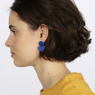 Frau mit kurzen braunen Haaren, die ein gelbes Oberteil und einen großen blauen geometrischen Ohrring trägt, im Profil vor einem schlichten hellen Hintergrund dargestellt.