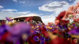 Modernes Gebäude mit geschwungenem Dach im Hintergrund, leuchtenden Blumen im Vordergrund und einem teilweise bewölkten Himmel darüber.