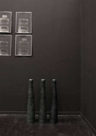 Vier abstrakte Schwarz-Weiß-Kunstwerke hängen an einer dunklen Wand über drei hohen, kegelförmigen schwarzen Skulpturen, die auf dem Boden stehen.