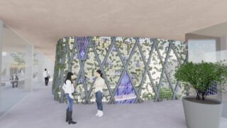 Zwei Personen stehen und unterhalten sich in der Nähe einer modernen, grün bepflanzten Wand mit Glasmuster in einem geräumigen, offenen Gebäude mit natürlichem Licht.