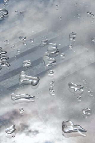 Auf einer reflektierenden Metalloberfläche verstreute Wassertropfen, auf denen schwache Reflexionen von Wolken und Himmel sichtbar sind.