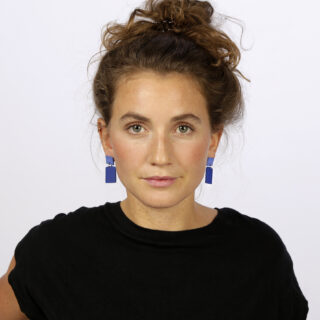 Eine Frau mit welligem braunem Haar in einem lockeren Dutt trägt ein schwarzes Oberteil und blaue geometrische Ohrringe und blickt vor einem schlichten weißen Hintergrund direkt in die Kamera.