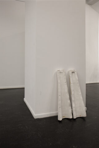 Zwei weiße Skulpturenobjekte lehnen an einer großen weißen Säule in einem Galerieraum mit dunklem Boden.