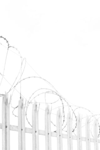 Vor einem strahlend weißen Himmel ist ein weißer Metallzaun mit aufgerolltem Stacheldraht oben drauf zu sehen.