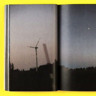 Das Foto eines aufgeschlagenen Buches zeigt eine Windkraftanlage in der Abenddämmerung mit einer dunklen Landschaft, einer schwachen Spiegelung der Anlage und einem einzelnen hellen Punkt am Himmel.