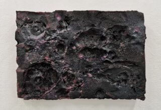 Ein rechteckiges, strukturiertes abstraktes Kunstwerk in Schwarz und Lila mit einer unebenen, kraterartigen Oberfläche ist an einer weißen Wand angebracht.