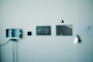 Im Vordergrund hängt an einem Faden eine kleine Metallglocke, im Hintergrund sind auf einer hellen Wand verschwommene Fotos oder Kunstwerke zu sehen.