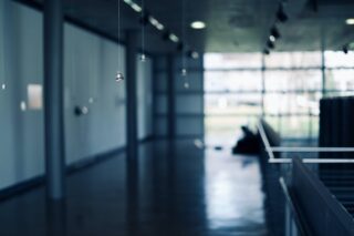 In einem leeren, modernen Gebäudekorridor mit großen Fenstern und polierten Böden hängen an dünnen Drähten kleine Metallgegenstände von der Decke.