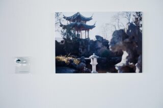 An einer weißen Wand hängt das Foto eines traditionellen asiatischen Pavillons, umgeben von Felsen und Bäumen. Links neben dem Foto befindet sich eine kleine transparente Gedenktafel.