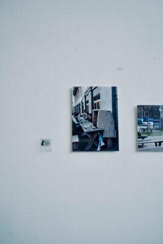 An einer hellen Wand hängt ein Foto, das eine Person zeigt, die mit einem großen Stapel Eierkartons an einem Tisch sitzt, wodurch ihr Gesicht teilweise verdeckt wird. Ein weiteres Foto und ein Etikett hängen ebenfalls an der Wand.