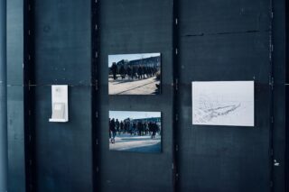 Auf einer dunklen Wand sind zwei Fotos von Menschen zu sehen, die in einer Gruppe spazieren gehen, die Skizze eines Auditoriums und eine kleine Papierprobe.