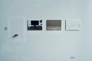 Drei gerahmte Bilder und ein kleines Objekt hängen an einer schlichten weißen Wand. Zu sehen sind ein Computertisch, eine verschwommene Szene und ein schwaches Diagramm. Darunter ist eine Steckdose zu sehen.