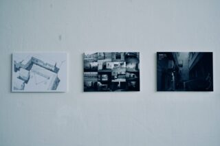 Drei rechteckige Fotografien oder Drucke werden in einer horizontalen Reihe auf einer schlichten weißen Wand angezeigt; jede zeigt architektonische oder urbane Bilder in monochromen Tönen.