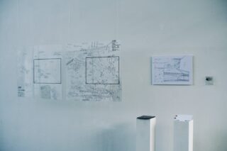 An einer Wand hängen zwei Transparentfolien mit technischen Zeichnungen neben einem aufgespannten Blatt mit Diagrammen, darunter stehen zwei weiße Präsentationsständer.