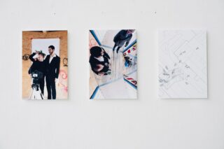 An einer weißen Wand hängen drei Kunstdrucke: ein Porträt von zwei Personen mit einer Kamera, eine Draufsicht von Menschen in einem verspiegelten Raum und eine abstrakte Zeichnung mit Linien und kleinen Formen.