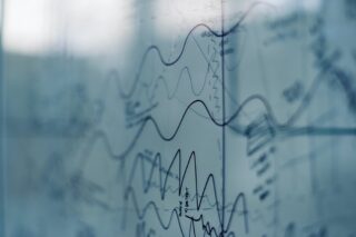 Auf einer transparenten Glastafel sind handgezeichnete Diagramme und Wellenlinien sichtbar, die verschiedene überlappende Datentrends vor einem unscharfen Hintergrund zeigen.