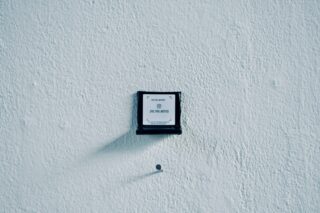 An einer strukturierten weißen Wand ist über einem einzelnen Nagel ein kleines Schild mit schwarzem Rahmen angebracht. Auf dem Schild steht „ZU IHRER BENACHRICHTIGUNG.“.