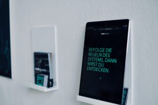 An einer weißen Wand ist ein Tablet angebracht, auf dem deutscher Text angezeigt wird. In der Nähe liegen auf einem Regal Zeitschriften und eine gefaltete Zeitung.