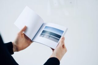 Eine Person hält ein Buch in der Hand und blättert darin. Auf einer Seite ist ein Foto eines ruhigen Ozeans mit einem Geländer im Vordergrund zu sehen.