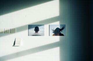 An einer sonnenbeschienenen Wand sind zwei Fotos zu sehen; eines zeigt den Rücken einer Person mit rasiertem Kopf, das andere eine Person mit seitlich wehendem Haar.