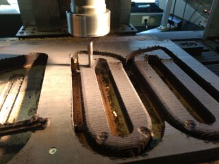 Eine CNC-Fräsmaschine schneidet Rillen in einen Materialblock. Im Arbeitsbereich sind Staub und Schmutz sichtbar.