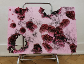 Ein rosa Brett mit schwarzen und dunkelroten Brandflecken und Löchern steht auf einem Metallgestell vor einer weißen Wand und einem Holzboden.