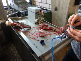 Eine Person verbindet Drähte und Komponenten auf einer Metallwerkbank und montiert oder testet ein elektronisches Gerät mit roten Kabeln und Leiterplatten.