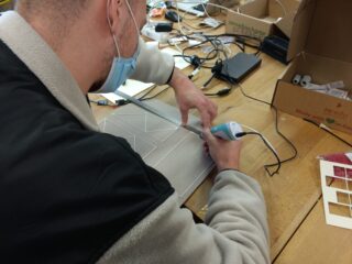 Eine Person mit einer Maske zeichnet an einem unaufgeräumten Arbeitsplatz mit einem Handgerät geometrische Formen auf ein Stück Pappe.