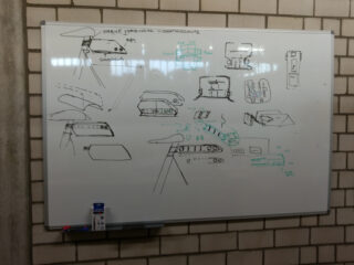 Whiteboard mit Skizzen und technischen Zeichnungen eines Gerätes, Schaltplänen, beschrifteten Bauteilen und Notizen in deutscher Sprache.