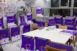 Ein Raum mit weiß gefliesten Wänden voller Graffiti, lila Plakaten mit der Aufschrift „RUN GANG“ auf Stühlen und Oberflächen und verstreuten Stühlen und Reinigungsmitteln um einen weißen Tisch herum.
