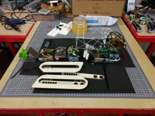 Eine überfüllte Werkbank mit Roboterkomponenten, Verkabelung, Leiterplatten, flügelähnlichen Holzteilen, Werkzeugen und Elektronik, angeordnet auf einer Schneidematte.