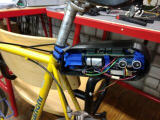 Eine Nahaufnahme eines gelben Fahrrads mit einem offenen elektronischen Gerät unter dem Sitz, das den Blick auf Leiterplatten, Kabel und Sensoren freigibt.