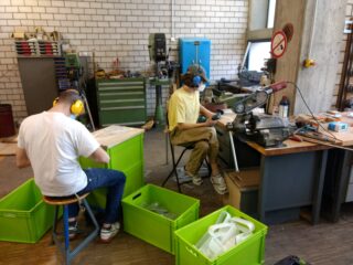 Zwei Personen in Schutzausrüstung arbeiten an getrennten Tischen in einer Werkstatt. Im Hintergrund sind Werkzeuge, grüne Lagerbehälter und verschiedene Geräte zu sehen.