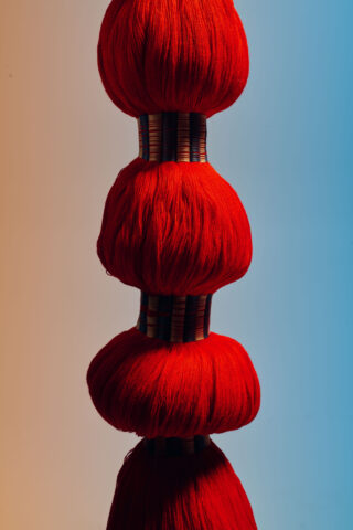 Vier Bündel leuchtend roten Garns sind vertikal auf zylindrischen Stäben vor einem Hintergrund mit Farbverlauf in Beige und Blau gestapelt.