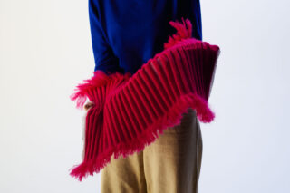 Eine Person in einem blauen Oberteil und einer beigen Hose hält einen strukturierten, gerippten, leuchtend rosa Stoff mit Fransenkanten vor einem hellen Hintergrund.