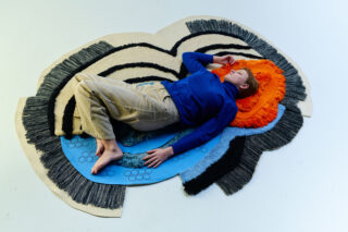 Eine Person in einem blauen Hemd und beigen Hosen liegt auf dem Rücken auf einem bunten, strukturierten Teppich mit abstrakten Mustern und verschiedenen Orange-, Blau-, Schwarz- und Cremetönen.