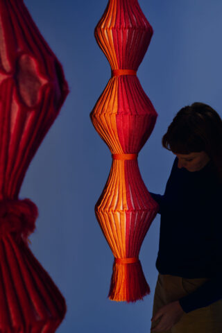 Eine Person steht neben einer großen, rot-orangenen Skulpturlampe mit plissierter Stoffstruktur vor blauem Hintergrund.