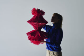 Eine Person in einem blauen Rollkragenpullover richtet eine große rote, plissierte Hängelampe vor einem schlichten, hellen Hintergrund.