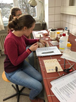 Zwei Personen sitzen an einer gefliesten Laborwerkbank, untersuchen Proben und machen sich Notizen auf Papier. In der Nähe liegen Laborgeräte und -zubehör.