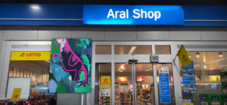 Der Eingang eines Supermarkts unter einem „Aral Shop“-Schild, mit einem farbenfrohen Dinosauriergemälde und Lotto-Schildern in der Nähe der Türen.
