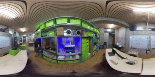 Ein Panoramablick auf einen Makerspace mit grünen Lagerbehältern, einem 3D-Drucker in einem blau beleuchteten Regal, Arbeitstischen, Computern und mit Skizzen bedeckten Whiteboards.
