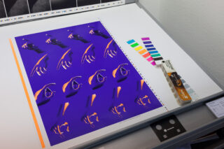 Ein Blatt mit wiederholten abstrakten Handgrafiken in Lila und Orange, Pantone-Farbmustern und einem Pantone-Farbführer auf einer weißen Oberfläche.